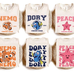 May include: Six cream-colored t-shirts featuring characters from "Finding Nemo." Designs include Nemo, Dory, and Peach, with text like "NEMO," "DORY," and "PEACH." Each shirt has "EST 2003" and "FINDING NEMO" printed on it. The shirts are arranged on woven mats.