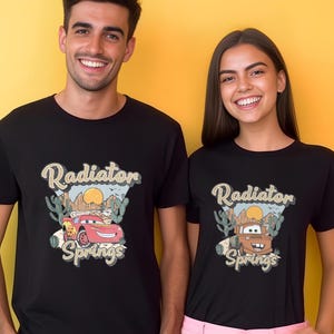 May include: Black t-shirts featuring a graphic of the Cars movie characters Lightning McQueen and Mater with the text "Radiator Springs". The design includes a desert landscape with cacti and a setting sun.
