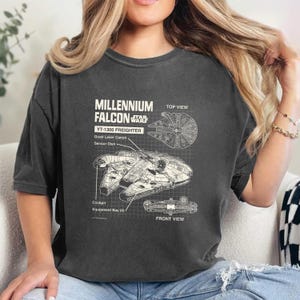 May include: A dark gray t-shirt with a white graphic of the Millennium Falcon spaceship from Star Wars. The graphic includes the text "Millennium Falcon", "YT-1300 Freighter", "Quad Laser Canon", "Sensor Dish", "Cockpit", "Equipment Bay (4)", "Top View", and "Front View".
