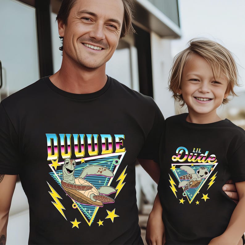May include: Black t-shirts with matching graphics. One shirt says "DUDE" in a retro font with a cartoon turtle, lightning bolts, and stars. The other shirt says "Lil Dude" with a similar design. A father and child are wearing the shirts.