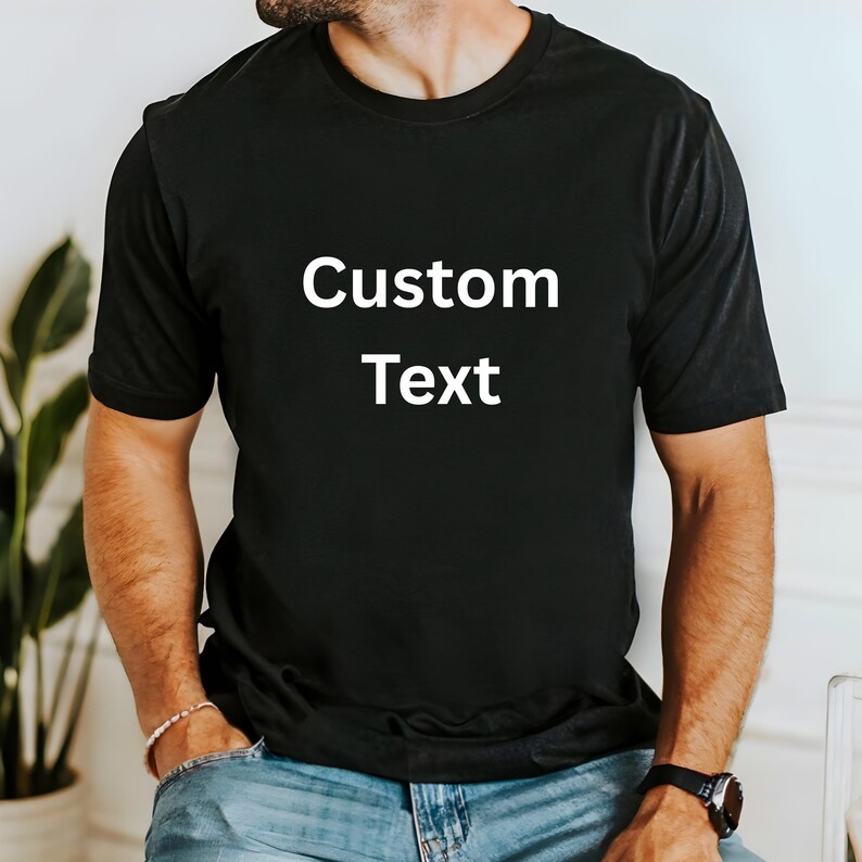 May include: Black crew neck t-shirt with the words "Custom Text" in white block letters. The shirt is a solid color and appears to be made of a soft material. The model is wearing blue jeans and a watch.