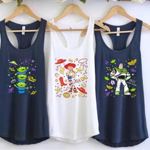 Three shirts hanging on a rack, each featuring a different character from the Toy Story franchise.