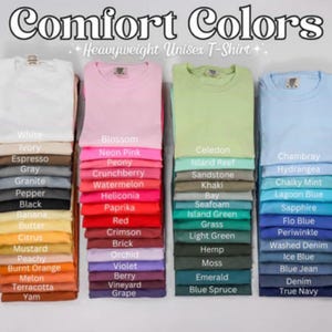 Może przedstawiać: Zdjęcie koszulek unisex Comfort Colors o dużej gramaturze w r&oacute;żnych kolorach, w tym białym, r&oacute;żowym, zielonym i niebieskim. Każda koszulka jest złożona i ułożona w stos, z paletą kolor&oacute;w pokazującą dostępne odcienie, takie jak "Blossom", "Chambray" i "True Navy."