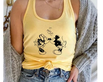 May include: Yellow tank top featuring a black and white graphic of Mickey and Minnie Mouse kissing, with three hearts above them. The top has a racerback design and a knotted hem. The model is wearing blue denim shorts and a gray cardigan.