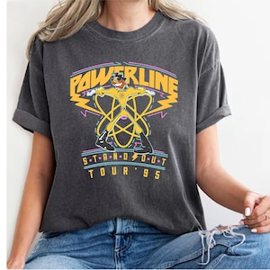 May include: A dark gray t-shirt with a vintage-style graphic of a cartoon character in a yellow and black suit. The graphic includes the text "POWERLINE" and "S.T.A.N.D.O.U.T TOUR '95".