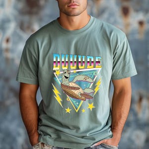 May include: Sage green t-shirt featuring a cartoon turtle with the word "DUUUUDE" in a retro font. The turtle is inside a yellow and blue triangle with lightning bolts and stars. The shirt is made of a soft material.
