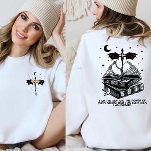 A woman wearing a white sweater with a graphic design on the back, including a bat, a book, and a crescent moon.