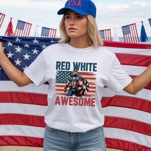 May include: A white short-sleeve t-shirt featuring a graphic of Captain America holding a shield in front of an American flag. The text "RED WHITE" appears in blue above the character, and "AWESOME" in red below, with a distressed vintage effect.