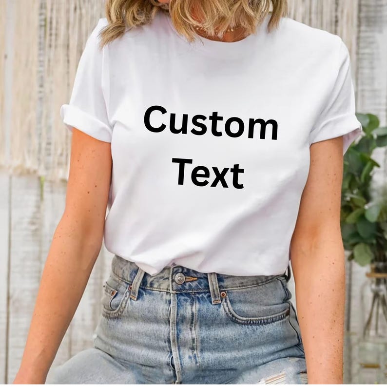 May include: White t-shirt with rolled sleeves and the words "Custom Text" printed in black. The shirt is paired with blue denim jeans. The overall style is casual and simple.