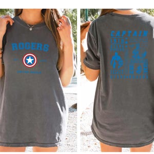 Puede incluir: Una camiseta gris con un logotipo azul y rojo de Capit&aacute;n Am&eacute;rica en la parte delantera y texto que dice "ROGERS" sobre el logotipo. La parte posterior de la camiseta tiene texto azul que dice "CAPTAIN" y "1918" con una estrella y "SUPER SOLDIER" con una estrella. El texto tambi&eacute;n dice "AVENGERS ASSEMBLE" y "ENDGAME" con una estrella.