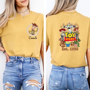 Puede incluir: Camiseta de color amarillo mostaza con un personaje de dibujos animados de Woody en un c&iacute;rculo a cuadros y el nombre "Charlie". La parte trasera de la camiseta muestra un gr&aacute;fico de "Toy Story" con personajes y el texto "Est. 1995".