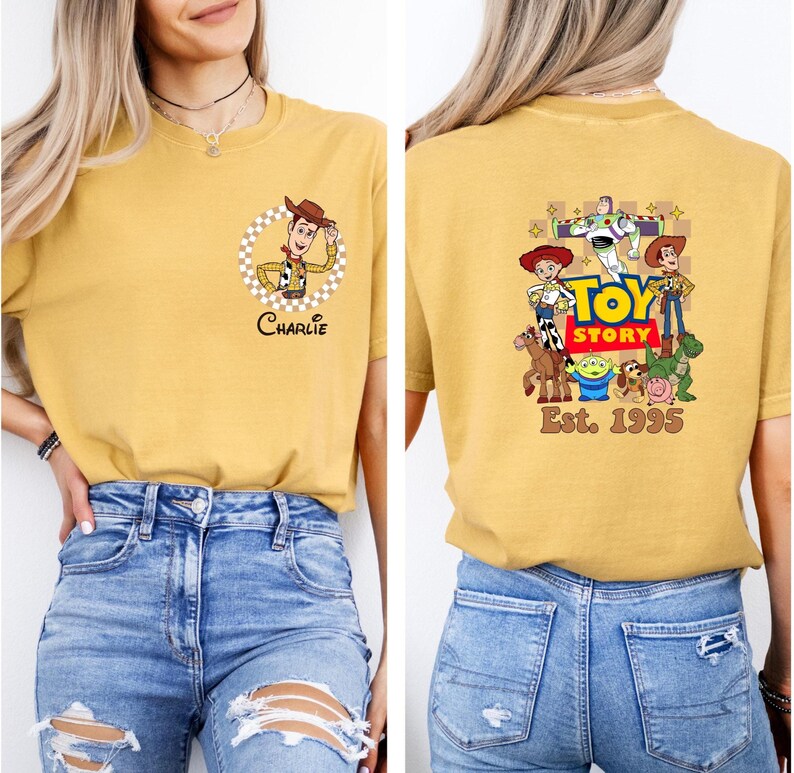 Puede incluir: Camiseta de color amarillo mostaza con un personaje de dibujos animados de Woody en un c&iacute;rculo a cuadros y el nombre "Charlie". La parte trasera de la camiseta muestra un gr&aacute;fico de "Toy Story" con personajes y el texto "Est. 1995".