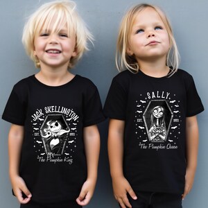 Pu&ograve; includere: Due bambini indossano magliette nere con grafiche bianche. Una maglietta dice "Jack Skellington" con un disegno a bara e "The Pumpkin King". L'altra dice "Sally" con un disegno a bara e "The Pumpkin Queen". Entrambe le magliette hanno "EST. 1993."