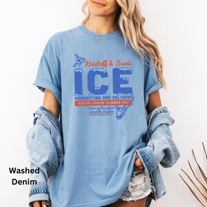 以下が含まれることがあります： クリストフとスヴェンのアイスハーベスティング＆デリバリーのグラフィックがプリントされた薄いブルーのTシャツ。グラフィックには、青と白のアイスロゴと、青と白で「Kristoff & Sven's ICE」というテキストが描かれています。「HARVESTING AND DELIVERY」というテキストは赤と白で書かれています。「WINTER-SPRING-SUMMER-FALL」というテキストは赤と白で書かれています。「Delivering throughout Arendelle Kingdom」というテキストは赤と白で書かれています。グラフィックには、クリストフとスヴェンのカートゥーン画像も含まれています。