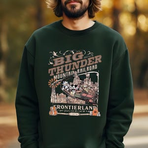 May include: Dark green sweatshirt featuring a graphic of the Big Thunder Mountain Railroad. The design includes the text "Big Thunder Mountain Railroad Frontierland Join Our Crew 1979" with cartoon characters and a train. The sweatshirt is a crew neck.