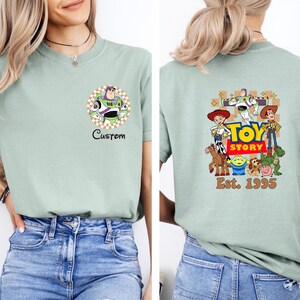 Puede incluir: Camiseta verde claro con un gr&aacute;fico de Buzz Lightyear en la parte delantera y un gr&aacute;fico de Toy Story en la parte trasera. El gr&aacute;fico frontal incluye la palabra "Custom". El gr&aacute;fico trasero incluye el texto "Toy Story" y "Est. 1995".