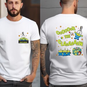Pu&ograve; includere: T-shirt bianca con un personaggio alieno verde che tiene una macchina a graffe con il testo "OOOOHHH THE CLAAAAW" e stelle sul retro. La parte anteriore della maglietta ha un'immagine pi&ugrave; piccola della macchina a graffe con il testo "EST. 1995".