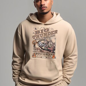 May include: Beige hoodie featuring a graphic of the Big Thunder Mountain Railroad ride. The design includes the ride's name, a train, and cartoon characters. The hoodie has a kangaroo pocket and a drawstring hood.