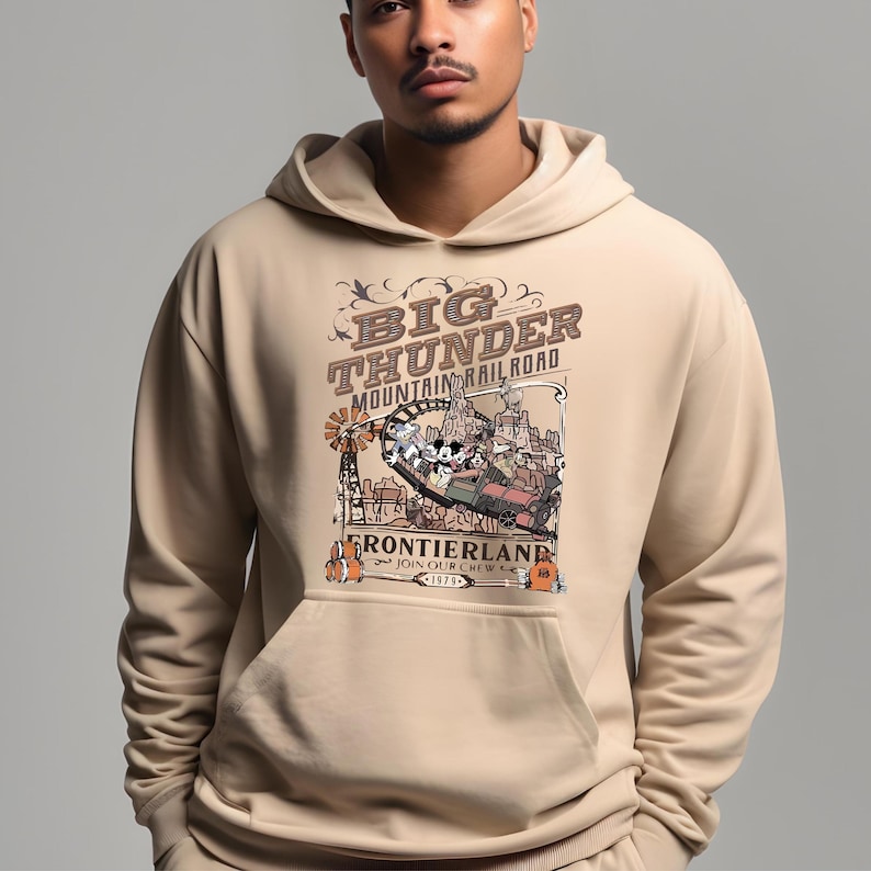 May include: Beige hoodie featuring a graphic of the Big Thunder Mountain Railroad ride. The design includes the ride's name, a train, and cartoon characters. The hoodie has a kangaroo pocket and a drawstring hood.