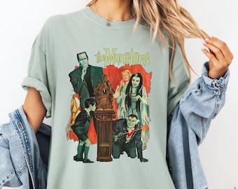 May include: A light green t-shirt featuring a graphic of the Munster family with the text "The Munsters" in green. The graphic includes images of Herman, Lily, Grandpa, Eddie, and Marilyn Munster. The shirt is a relaxed fit.