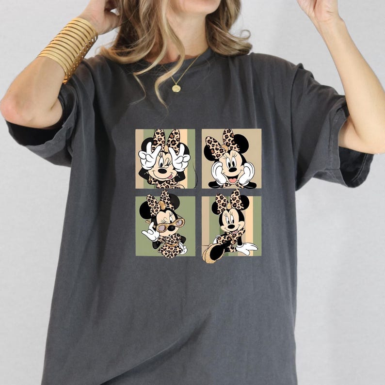 May include: Dark grey t-shirt with a four-panel design of Minnie Mouse in different poses. Each panel shows Minnie in a leopard print bow and outfit, set against a beige and green striped background. The t-shirt has a casual, oversized fit.