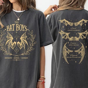 May include: A dark gray t-shirt with a gold graphic print of bats and the text "THE BAT BOYS.. Rhysand Azriel Cassian".