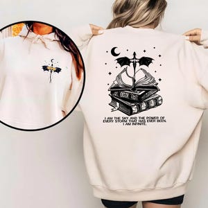 A person wearing a white sweatshirt with a black graphic design on the back, which features a dragon, books, and a crescent moon.