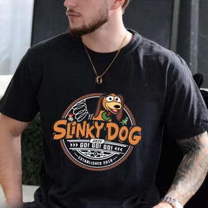 Może przedstawiać: Czarny t-shirt z okrągłą grafiką z napisem "Slinky Dog" w kolorze pomarańczowym i białym. Grafika zawiera ilustrację Slinky Dog, postaci z kresk&oacute;wki, oraz tekst "GO! GO! GO!" i "Established 2018".