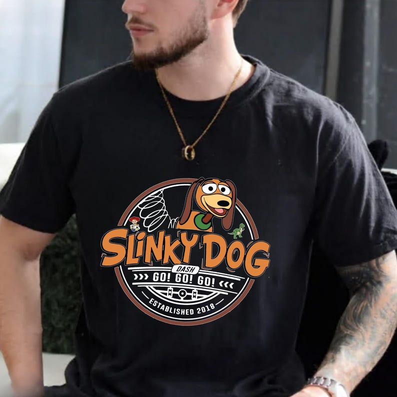 Może przedstawiać: Czarny t-shirt z okrągłą grafiką z napisem "Slinky Dog" w kolorze pomarańczowym i białym. Grafika zawiera ilustrację Slinky Dog, postaci z kresk&oacute;wki, oraz tekst "GO! GO! GO!" i "Established 2018".