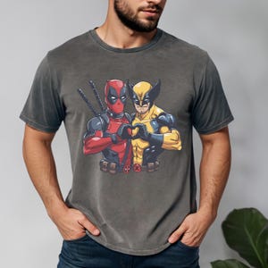 May include: Dark gray t-shirt featuring a graphic of Deadpool and Wolverine forming a heart with their hands. Deadpool is in a red and black suit, and Wolverine is in a yellow and blue suit. The shirt has a vintage wash.