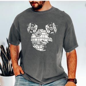 May include: Gray t-shirt featuring a white graphic design of the Death Star with TIE fighters as ears. The design is a parody of a famous cartoon character. The shirt is made of a soft, comfortable material.
