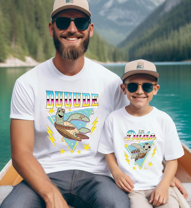 May include: Two white t-shirts with matching designs. The shirts feature the word "DUUUUDE" and "Lil Dude" in a retro font, along with a cartoon turtle. Both wear sunglasses and hats. The background shows a lake and mountains.