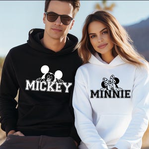 A man and a woman wearing matching Mickey Mouse-themed hoodies stand together against a desert landscape background.