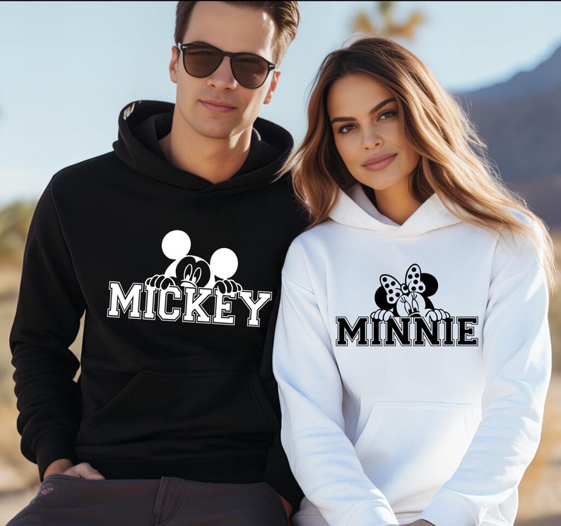 A man and a woman wearing matching Mickey Mouse-themed hoodies stand together against a desert landscape background.
