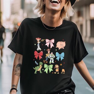 K&ouml;nnte beinhalten: Schwarzes T-Shirt mit einer farbenfrohen Grafik von Figuren und Schleifen aus dem Animationsfilm. Das Design umfasst Bilder eines Schweins, eines Dinosauriers, eines Hundes und anderer Charaktere. Das Shirt hat einen Rundhalsausschnitt und kurze &Auml;rmel.