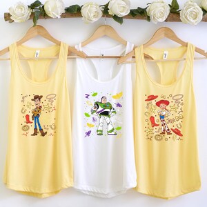 Three yellow tank tops hanging on a wooden hanger, each featuring a different character from the Toy Story franchise.