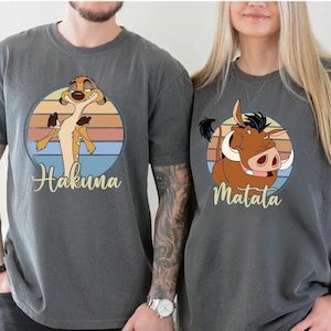 May include: Two gray t-shirts featuring characters from The Lion King. One shirt has Timon with "Hakuna" and the other has Pumbaa with "Matata" in a retro sunset circle design. The shirts are short-sleeved.