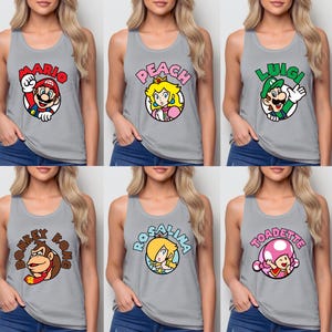 May include: A collection of six grey tank tops, each featuring a circular graphic of a Nintendo character. The characters include Mario, Peach, Luigi, Donkey Kong, Rosalina, and Toadette, with their names printed in colorful, bold lettering above.