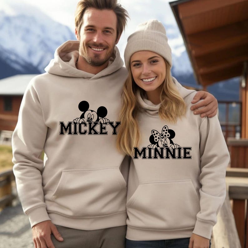 A man and a woman are standing together, both wearing matching Mickey Mouse-themed hoodies.