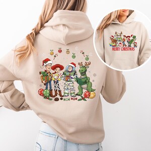 May include: Beige hoodie featuring a colorful graphic of animated characters in Santa hats, holding gifts and reading a book. The design includes Christmas ornaments and candy canes. The smaller image on the right says "MERRY CHRISTMAS" in red.