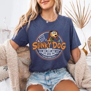 Może przedstawiać: Granatowa koszulka z logo Slinky Dog Dash. Logo zawiera ilustrację Slinky Dog, tekst "Slinky Dog Dash" oraz frazę "GO! GO! GO!". Koszulka ma okrągły dekolt i kr&oacute;tkie rękawy.