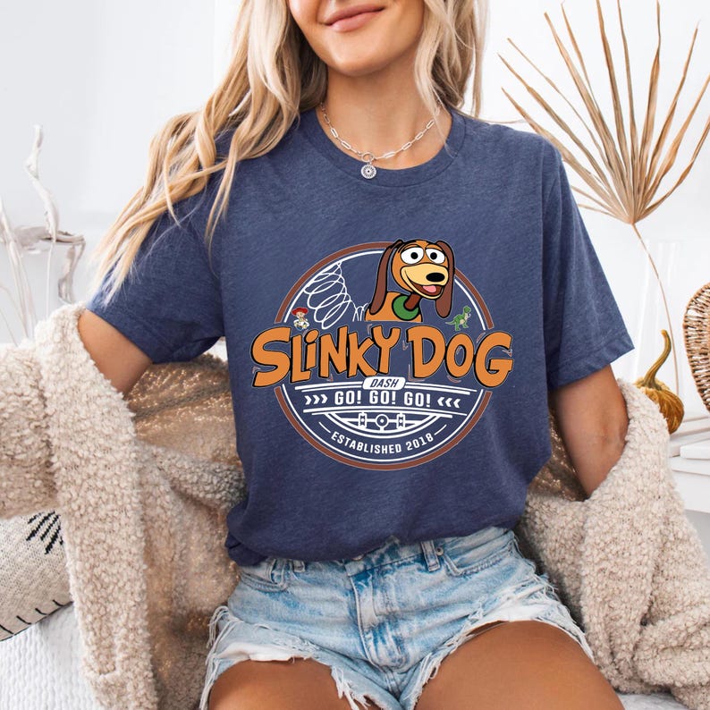 Może przedstawiać: Granatowa koszulka z logo Slinky Dog Dash. Logo zawiera ilustrację Slinky Dog, tekst "Slinky Dog Dash" oraz frazę "GO! GO! GO!". Koszulka ma okrągły dekolt i kr&oacute;tkie rękawy.