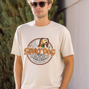 Może przedstawiać: Kremowa koszulka z okrągłą grafiką. Grafika zawiera napis "Slinky Dog Dash" oraz ilustrację Slinky Dog. Grafika zawiera r&oacute;wnież tekst "GO! GO! GO!" i "ESTABLISHED 2018".