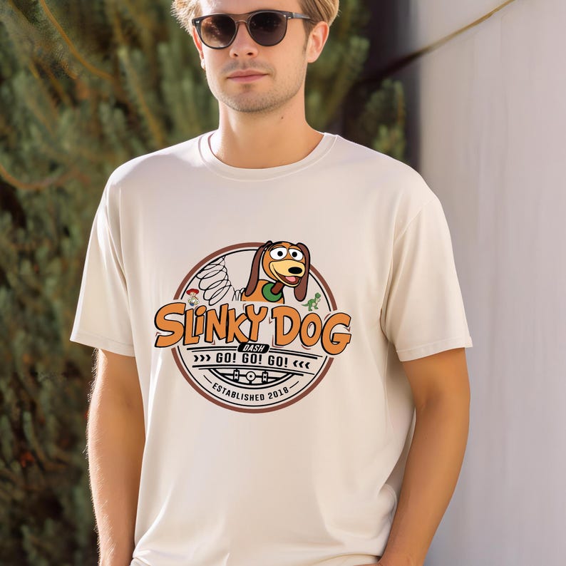 Może przedstawiać: Kremowa koszulka z okrągłą grafiką. Grafika zawiera napis "Slinky Dog Dash" oraz ilustrację Slinky Dog. Grafika zawiera r&oacute;wnież tekst "GO! GO! GO!" i "ESTABLISHED 2018".