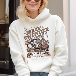 May include: Cream-colored hoodie featuring a graphic of the Big Thunder Mountain Railroad. The design includes the text "Big Thunder Mountain Railroad" and "Frontierland" with a cartoon train and characters. The hoodie has a front pocket and a drawstring hood.