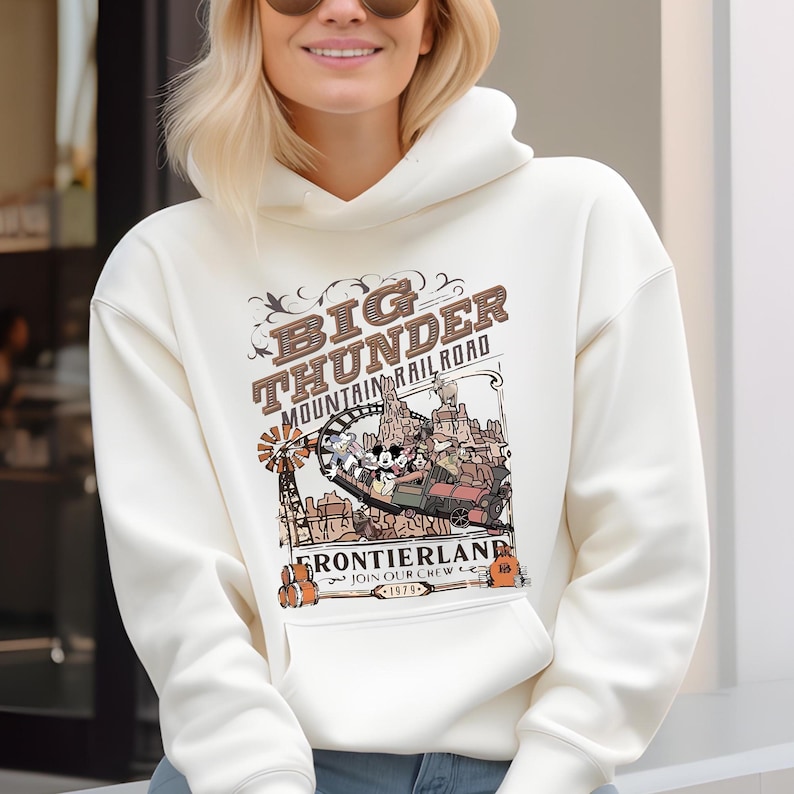 May include: Cream-colored hoodie featuring a graphic of the Big Thunder Mountain Railroad. The design includes the text "Big Thunder Mountain Railroad" and "Frontierland" with a cartoon train and characters. The hoodie has a front pocket and a drawstring hood.