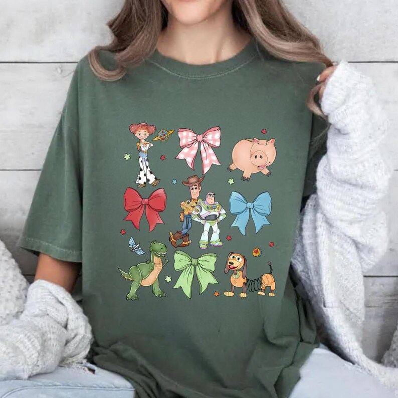 K&ouml;nnte beinhalten: Gr&uuml;nes T-Shirt mit Cartoon-Illustrationen von Figuren und Schleifen. Das Design umfasst ein rosa Schwein, einen gr&uuml;nen Dinosaurier und einen braunen Hund. Weitere Charaktere sind ein Cowgirl, ein Cowboy und ein Weltraumranger.