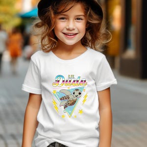 May include: White t-shirt featuring a cartoon turtle graphic with the text "Lil Dude" in a colorful, retro font. The turtle is inside a blue and green triangle with yellow stars and lightning bolts.