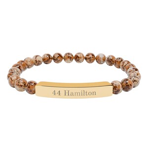 Peut inclure: Un bracelet de perles avec des perles tachetées marron et beige. Une barre rectangulaire dorée au centre est gravée du texte "44 Hamilton". Le bracelet est conçu pour être porté au poignet.