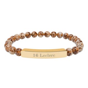 Könnte beinhalten: Armband mit braunen und beigefarbenen, gesprenkelten Perlen. Ein goldfarbener, rechteckiger Steg in der Mitte ist mit dem Text "16 Leclerc" graviert. Das Armband ist auf einem Gummiband aufgezogen.